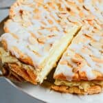 A slice of almond-topped, iced Gluten Free Almond Eclair Cake is being lifted from a larger round cake on a plate.