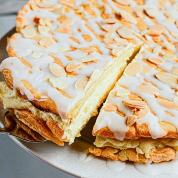 A slice of almond-topped, iced Gluten Free Almond Eclair Cake is being lifted from a larger round cake on a plate.