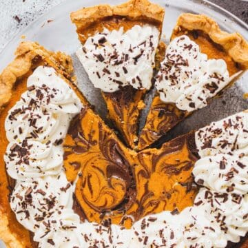 Pumpkin pie with chocolate swirls, topped with whipped cream and chocolate shavings, on a glass plate.