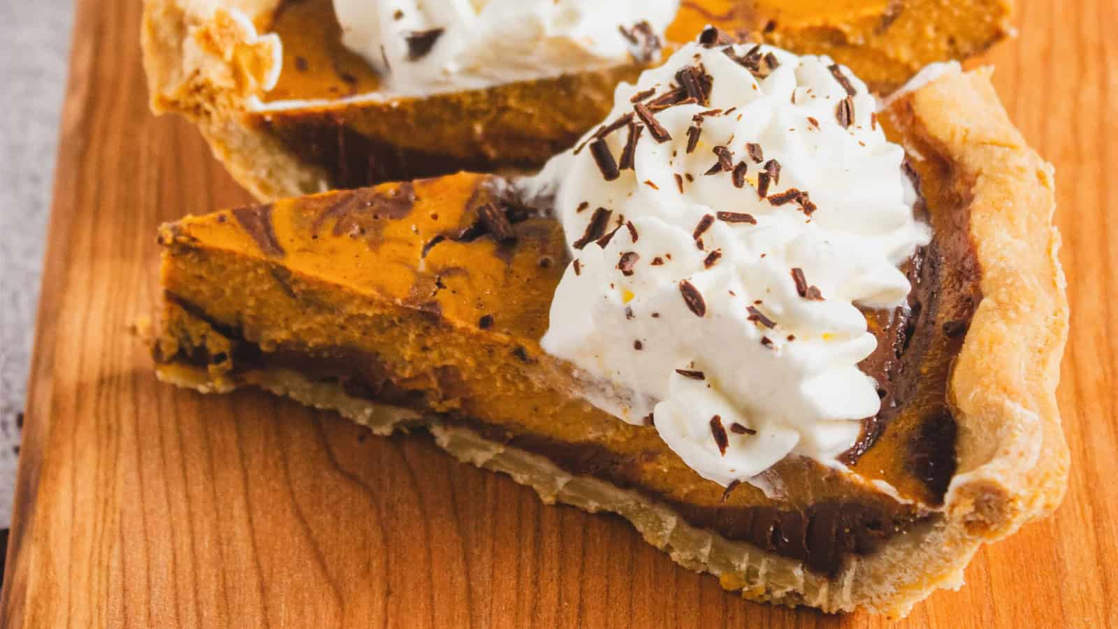 A slice of pumpkin pie with whipped cream and chocolate shavings on a wooden surface.