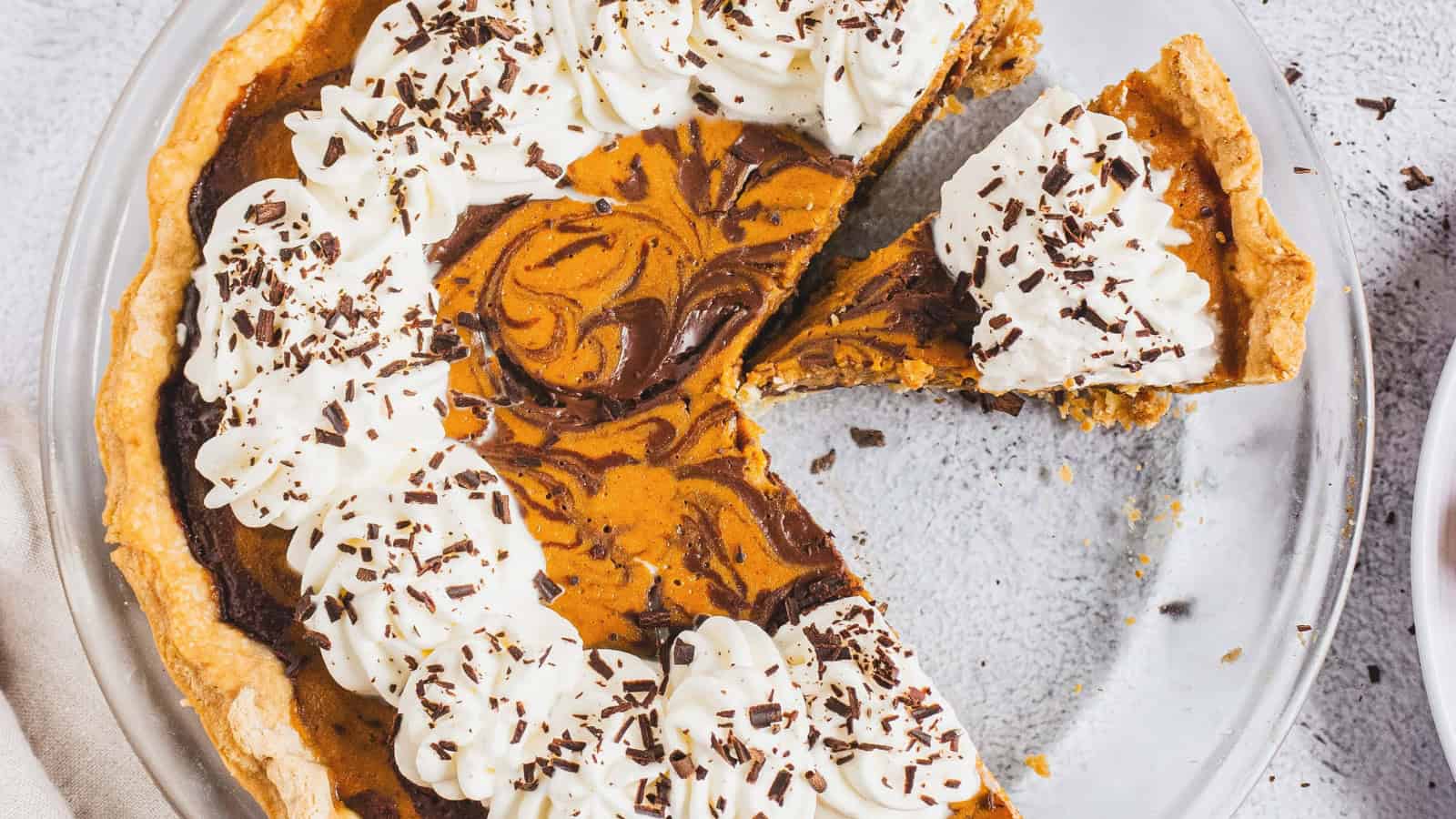 Pumpkin pie with chocolate swirl, topped with whipped cream and chocolate shavings, on a glass plate.