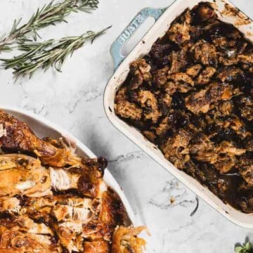 Roasted turkey pieces and stuffing in dishes on a marble surface with herbs nearby.