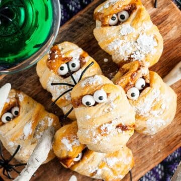 Pastry treats decorated as mummies with candy eyes on a wooden board, next to a green drink and fake white bones.