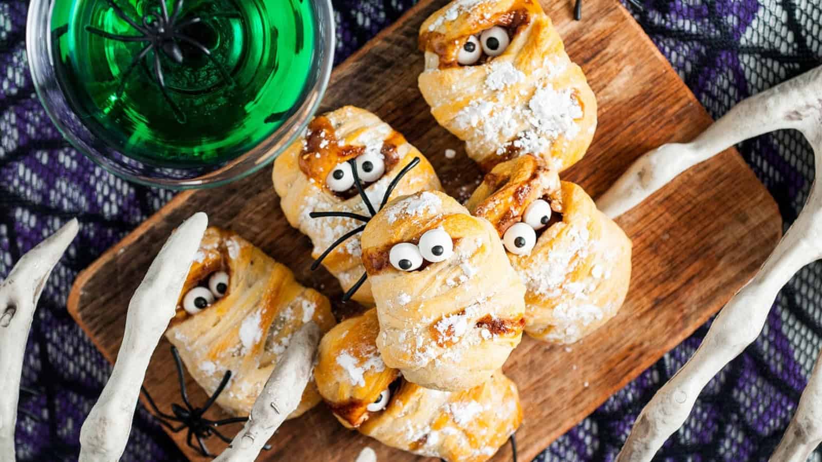 Pastry treats decorated as mummies with candy eyes on a wooden board, next to a green drink and fake white bones.