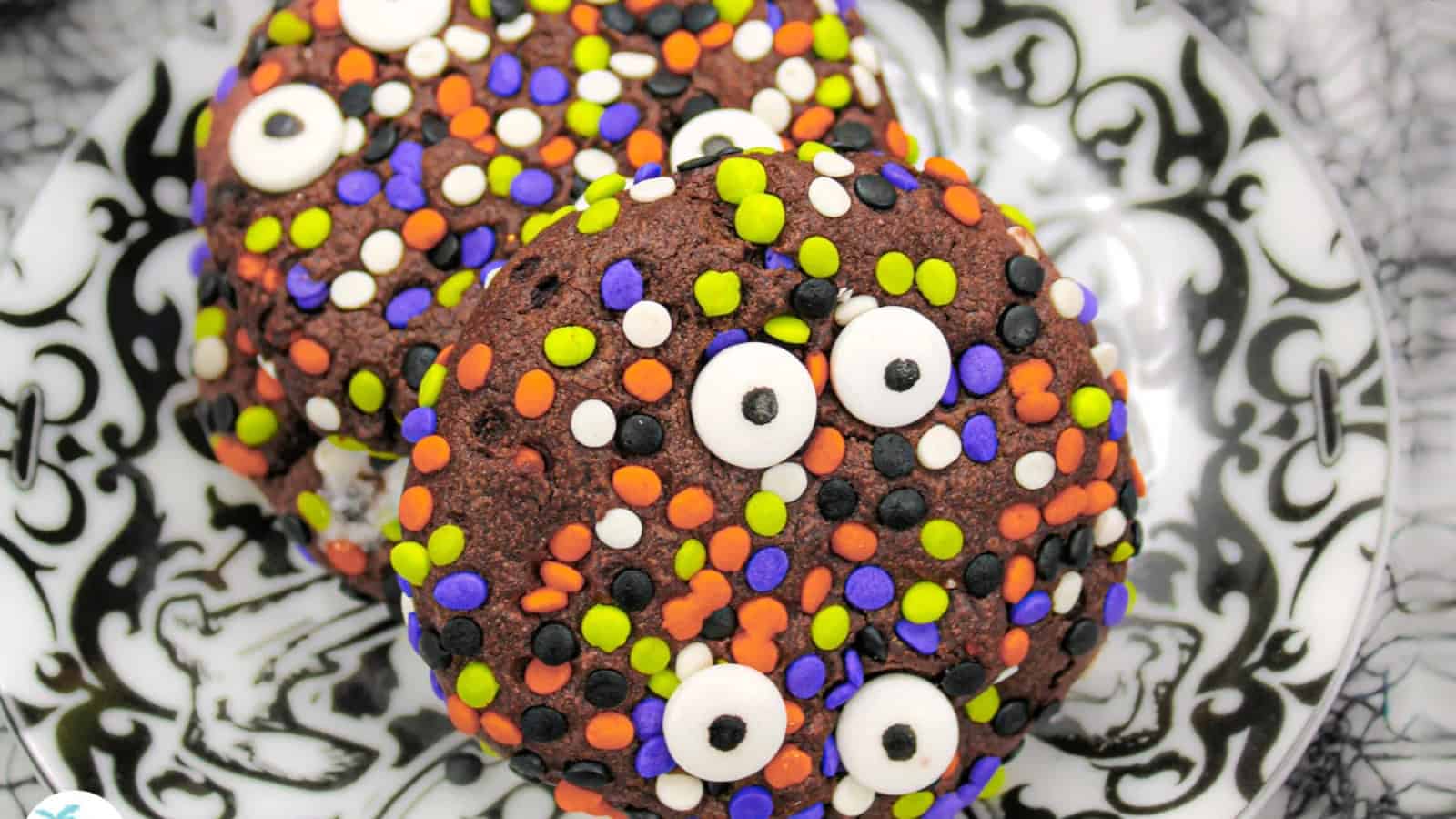 A stack of three chocolate cookies covered in colorful sprinkles, including orange, purple, green, white, and black, with candy eyes. They are on a decorative black and white plate with a patterned cloth in the background.