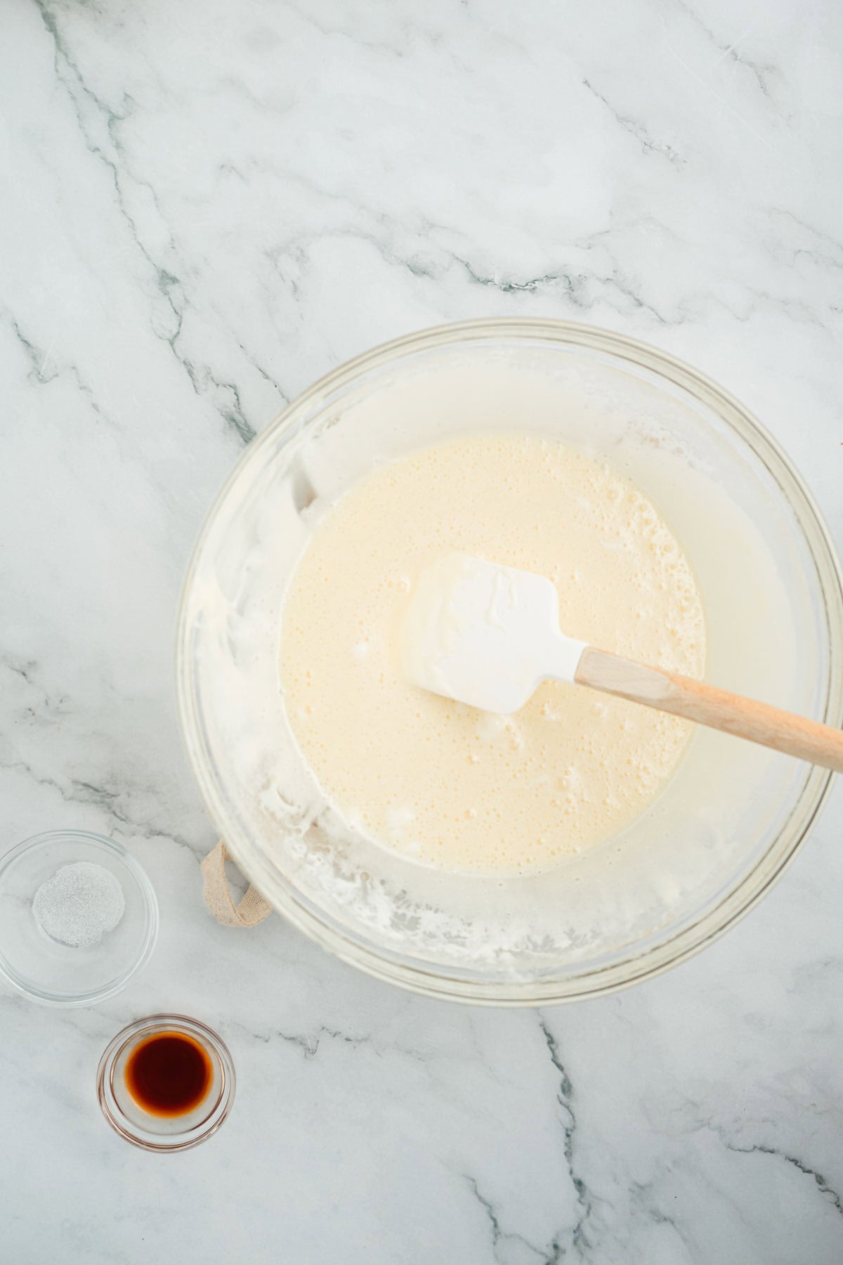 Glass bowl with whisked batter and a spatula, next to small bowls of salt and vanilla on a marble surface—perfect for whipping up Halloween Candy Rice Krispies treats.
