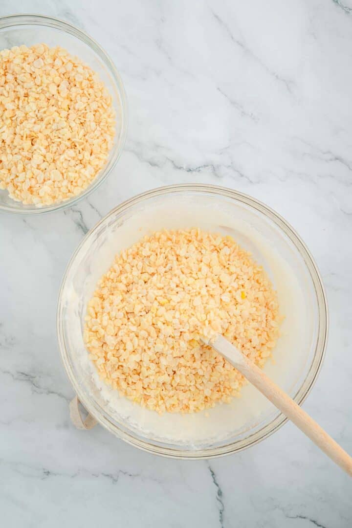Two glass bowls with crispy rice cereal, one mixed with melted marshmallow for Halloween Candy Rice Krispies, on a marble countertop.