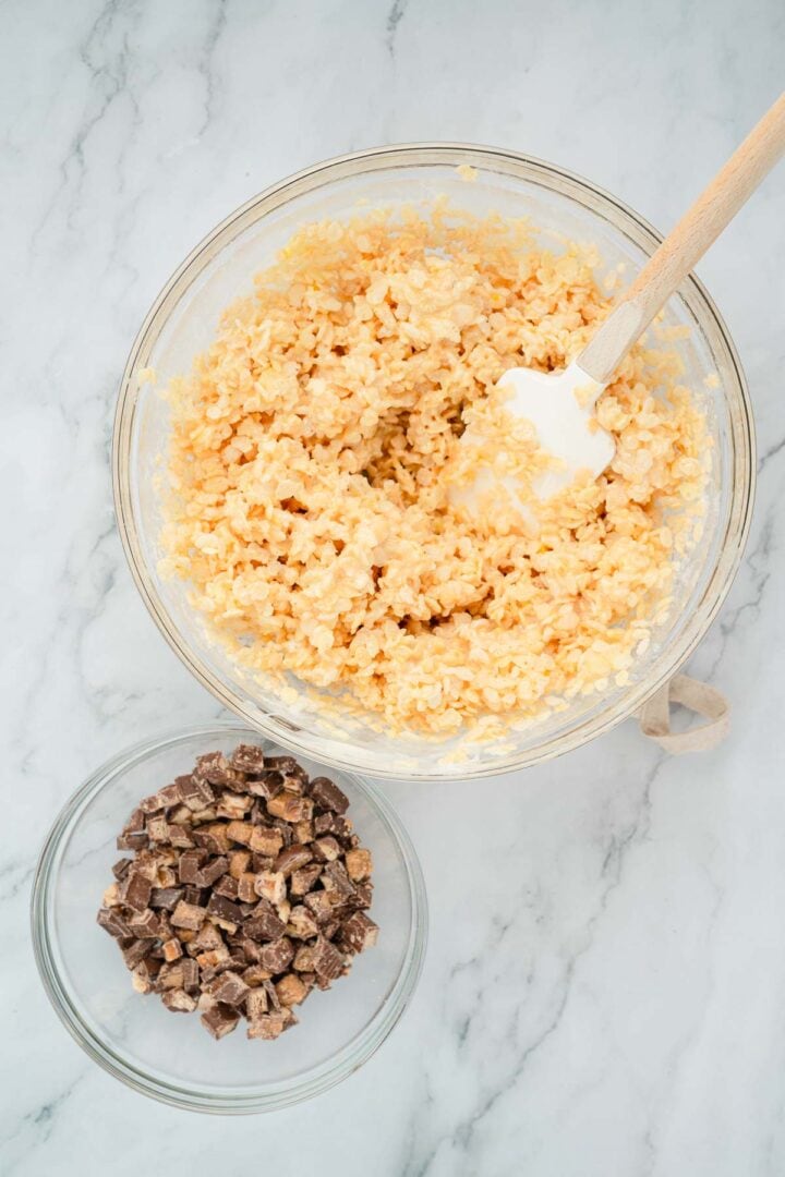 A bowl of Halloween Candy Rice Krispies mixture sits with a spatula beside a bowl of chopped chocolate candy on a marble surface.