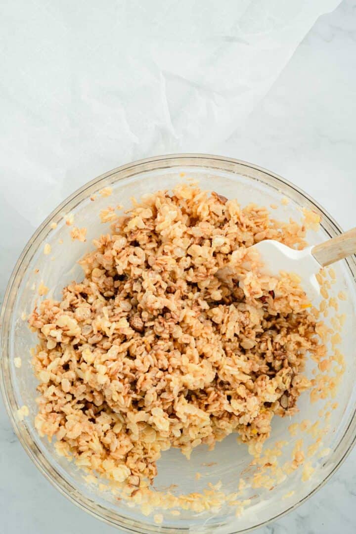 A glass bowl of Halloween Candy Rice Krispies treat mixture is being stirred with a white spatula on a marble surface.