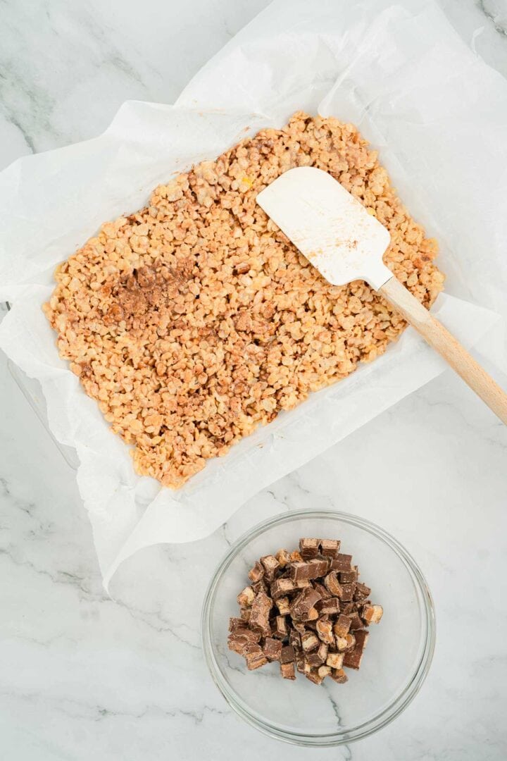 A pan of Halloween Candy Rice Krispies sits with a spatula and a bowl of chopped chocolate candies on a marble surface.