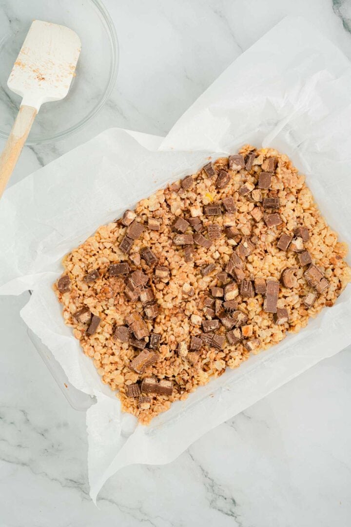 Halloween Candy Rice Krispies treats with chocolate chunks in a baking dish lined with parchment paper, next to a spatula.