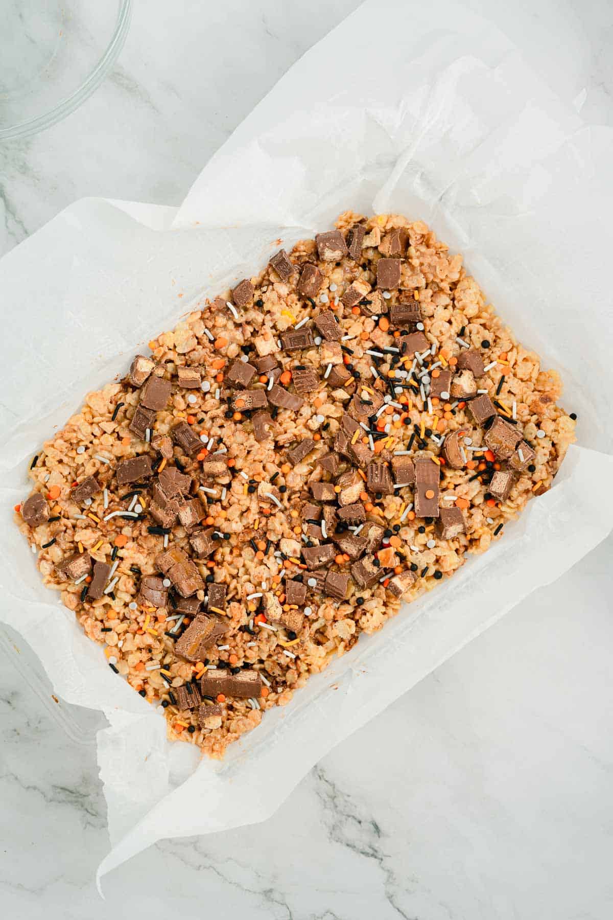 A pan of Halloween Candy Rice Krispies treats topped with chocolate chunks and colorful sprinkles on parchment paper.