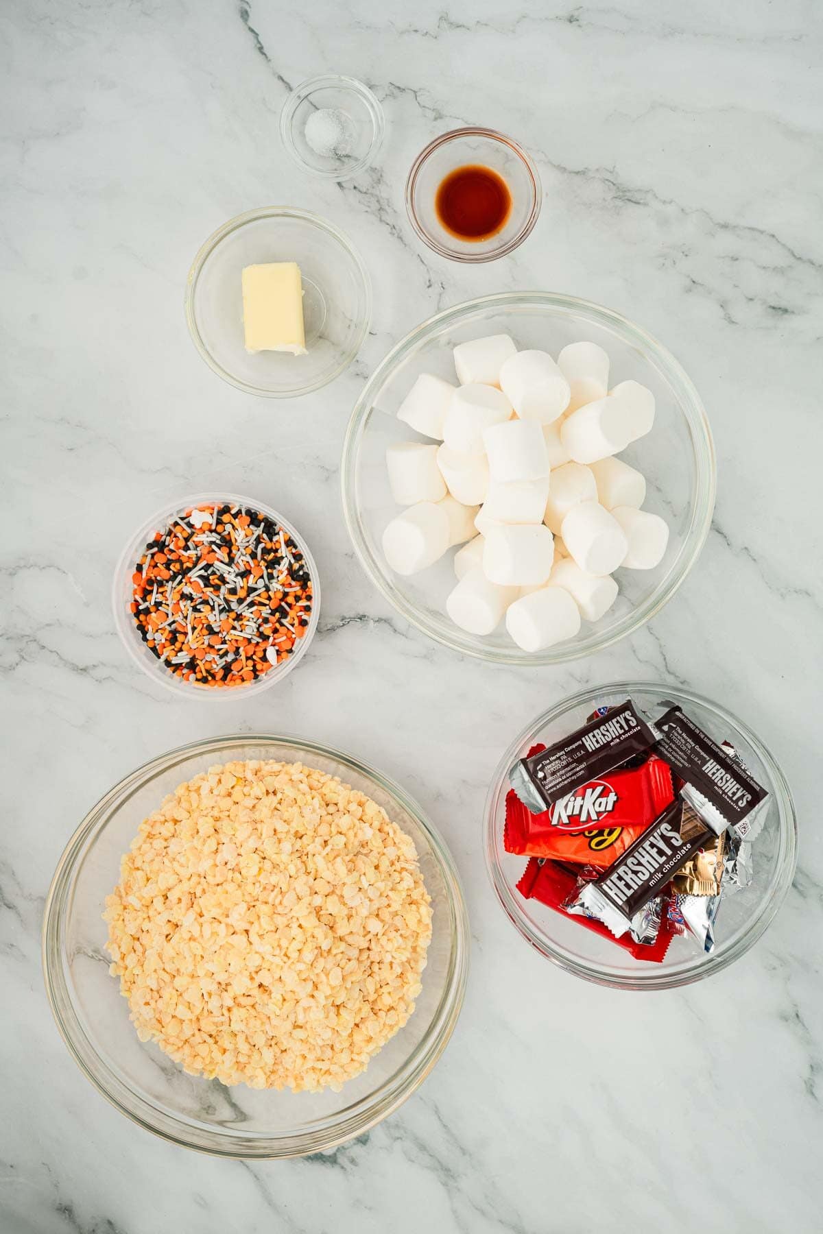 Bowls of marshmallows, rice cereal, Halloween sprinkles, candy bars, butter, and vanilla sit on a marble surface—everything you need to create festive Halloween Candy Rice Krispies treats.
