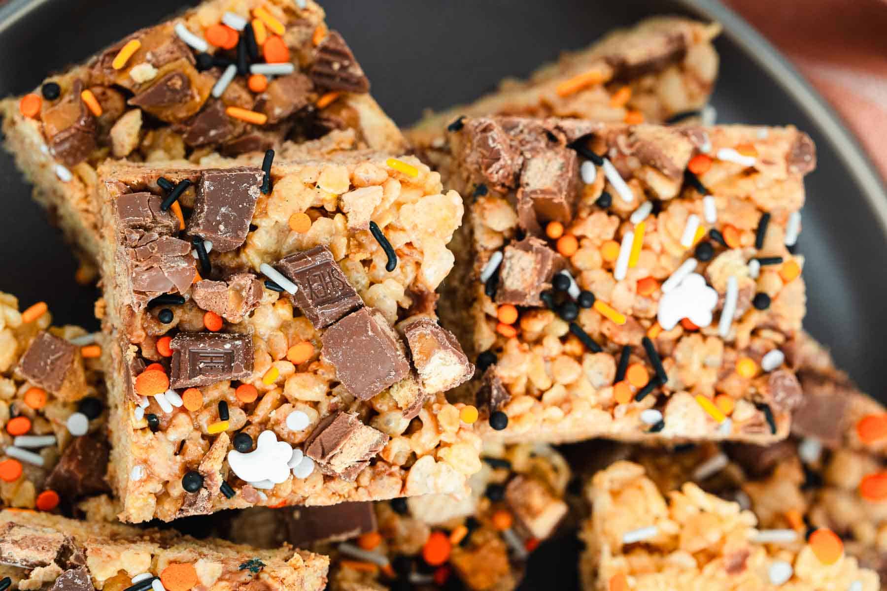 Halloween Candy Rice Krispies treats topped with festive sprinkles and chopped chocolate candy sit on a dark plate, perfect for spooky celebrations.