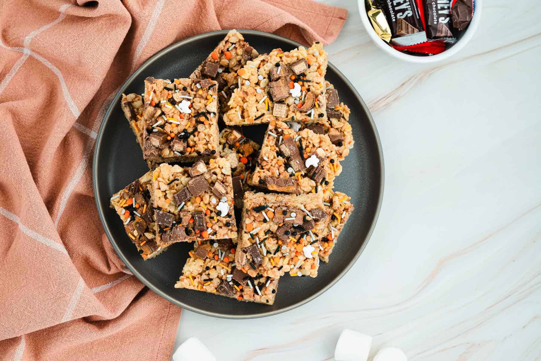 A plate of Halloween Candy Rice Krispies treats, topped with chocolate and sprinkles, sits next to a pink cloth and a bowl of festive candy.