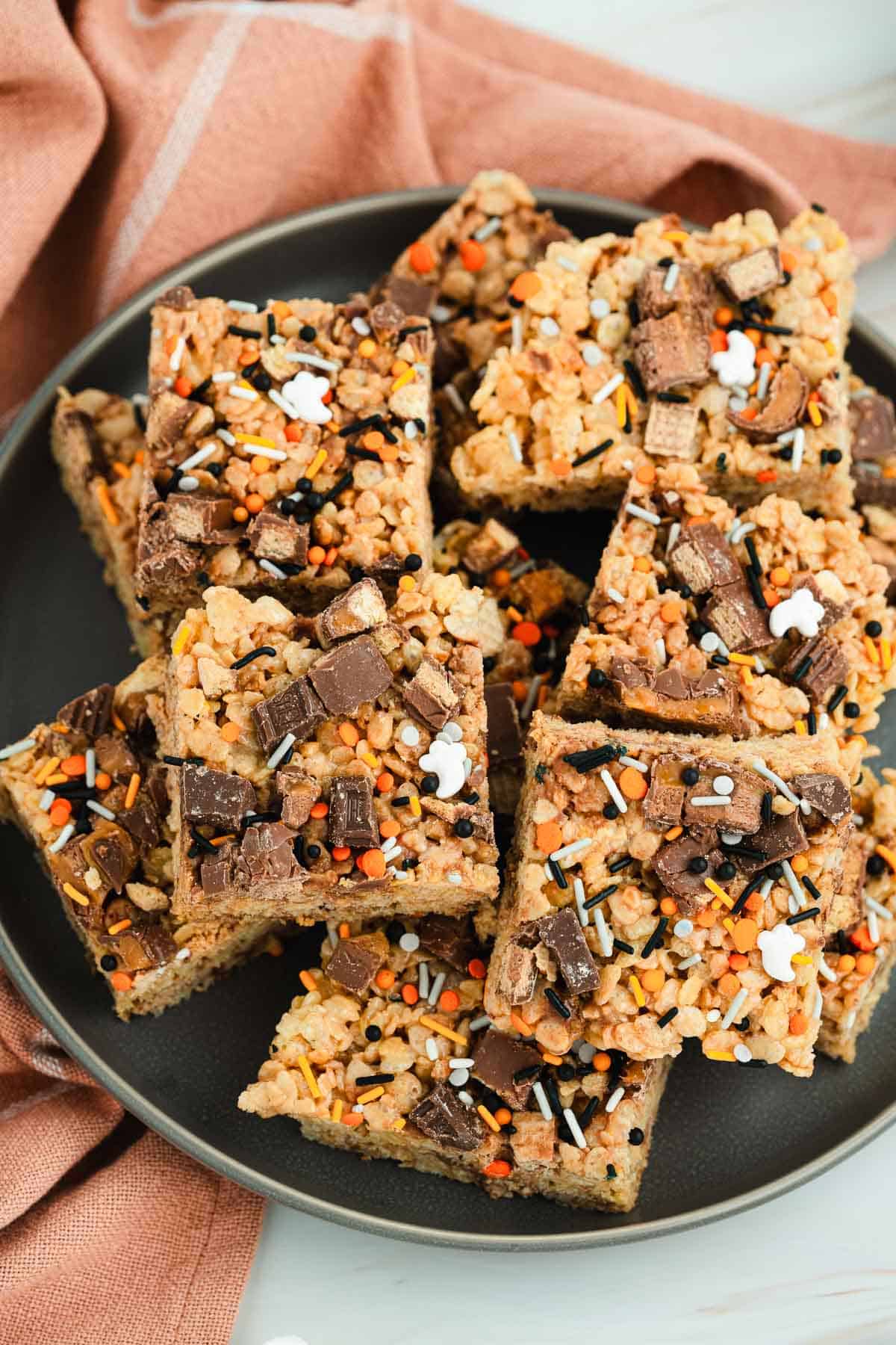 A plate of Halloween Candy Rice Krispies treats topped with chocolate pieces and colorful orange, black, and white sprinkles.