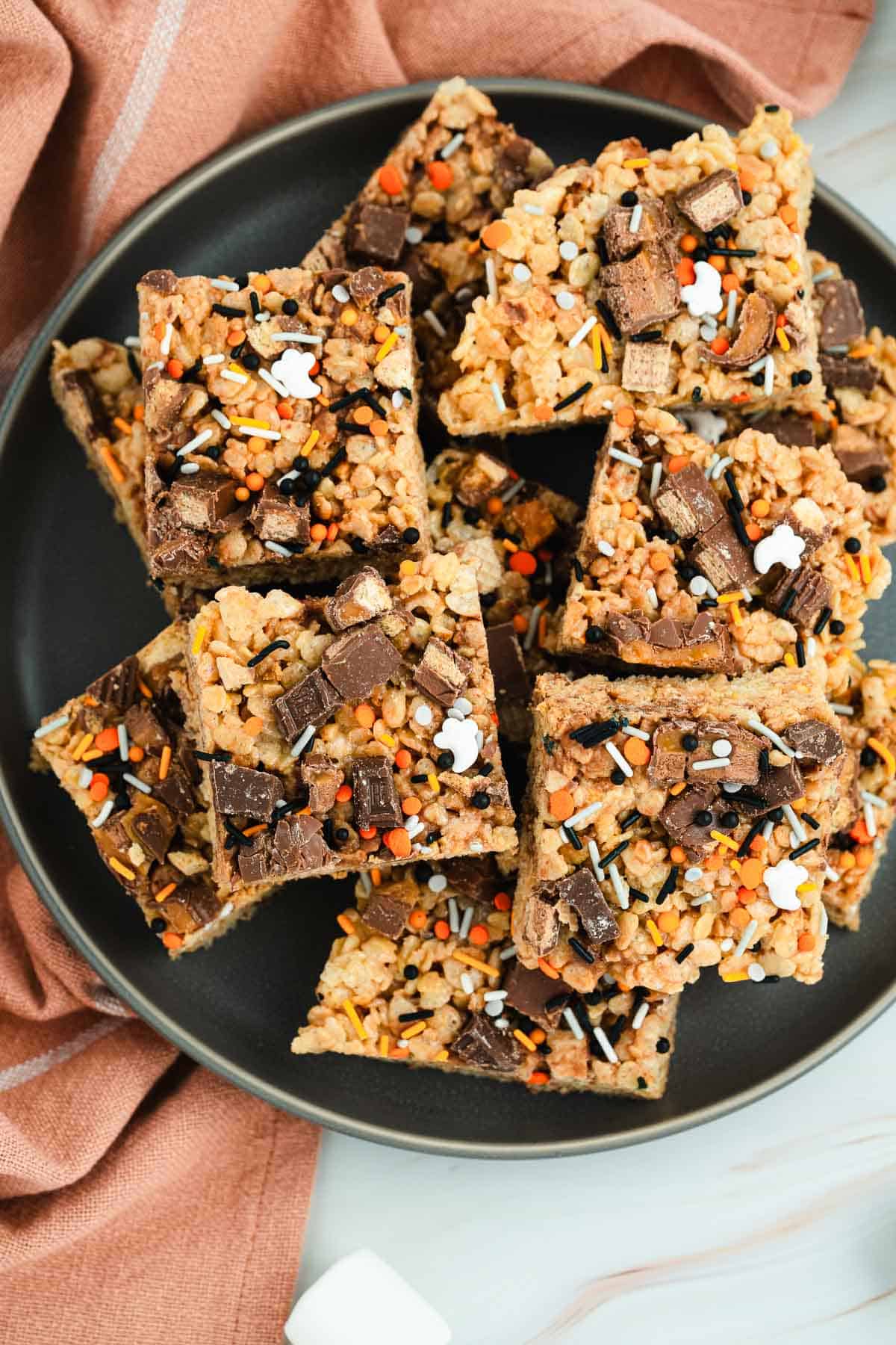 Halloween Candy Rice Krispies treats topped with chocolate pieces and festive Halloween sprinkles, stacked on a black plate.