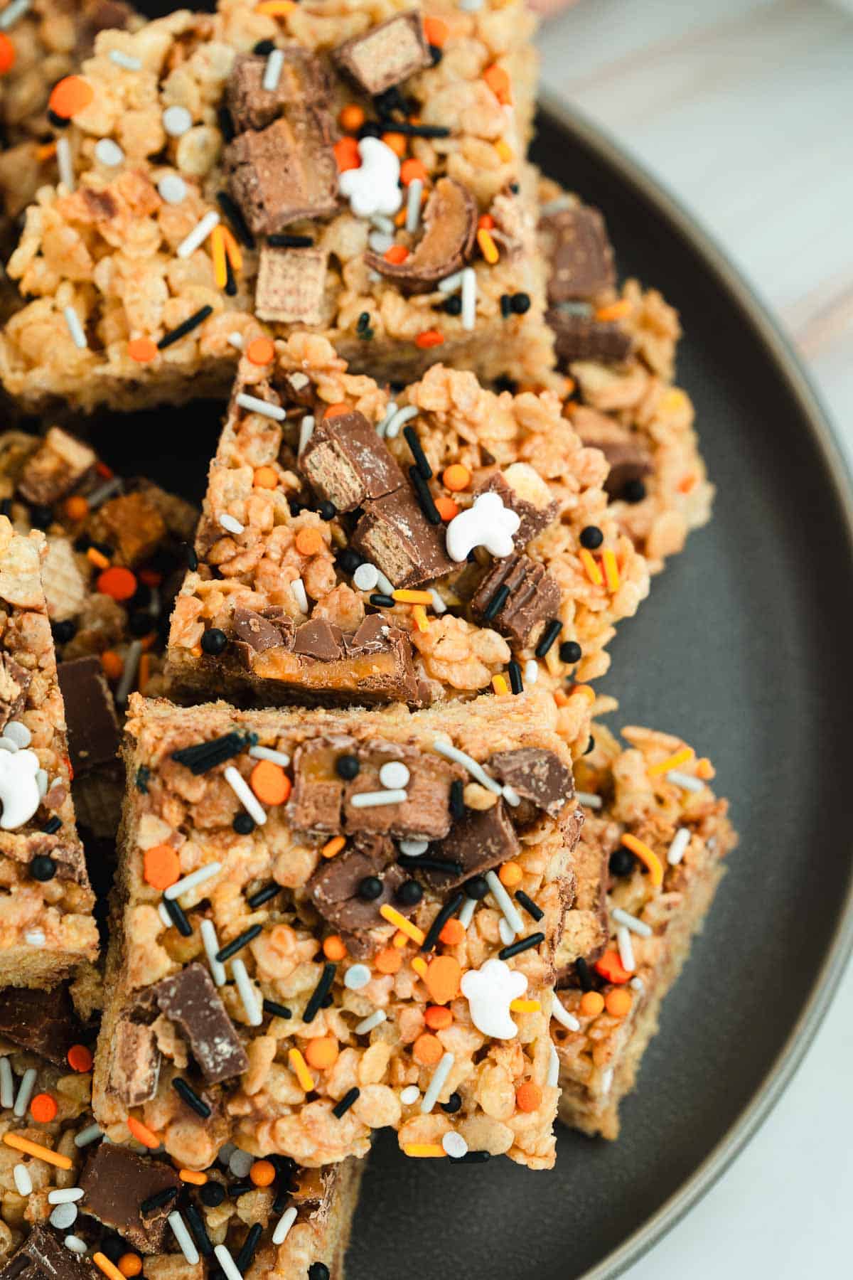 Halloween Candy Rice Krispies treats topped with chocolate chunks and colorful Halloween-themed sprinkles are served on a dark plate.
