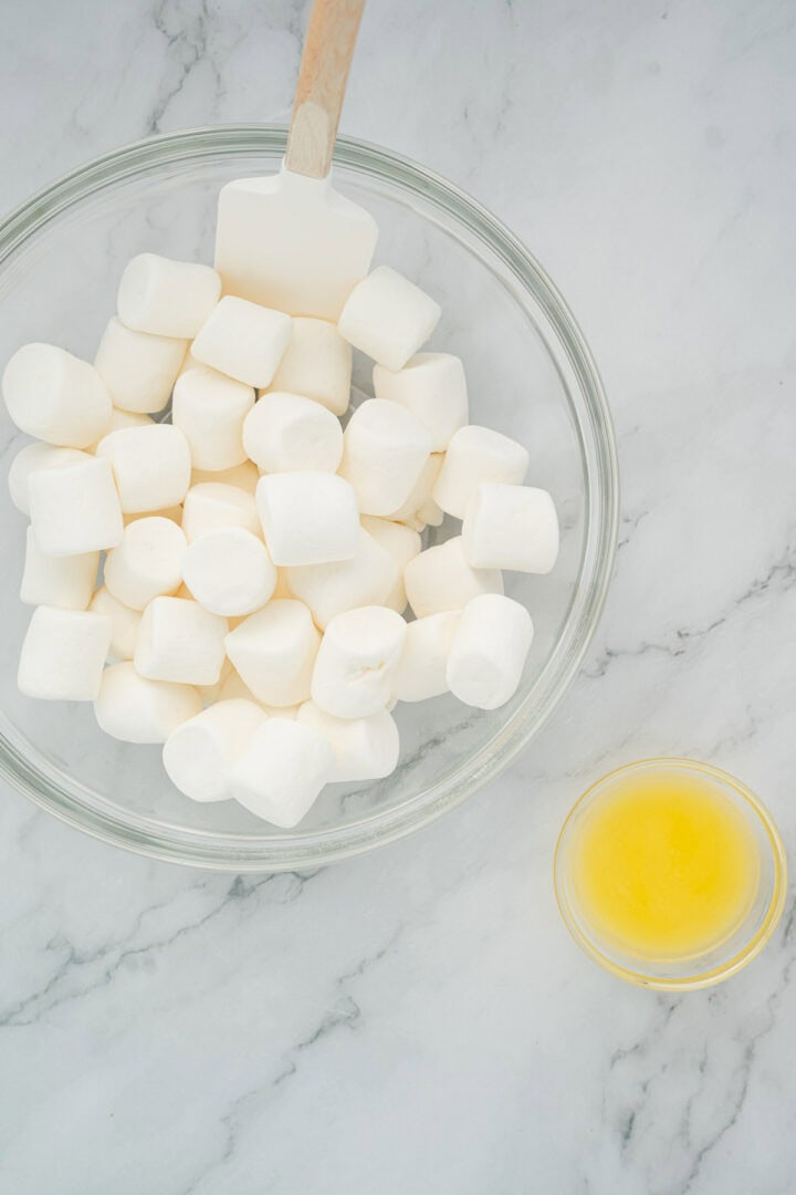 A glass bowl of marshmallows and a small bowl of melted butter on a marble countertop—perfect for making Halloween Candy Rice Krispies.