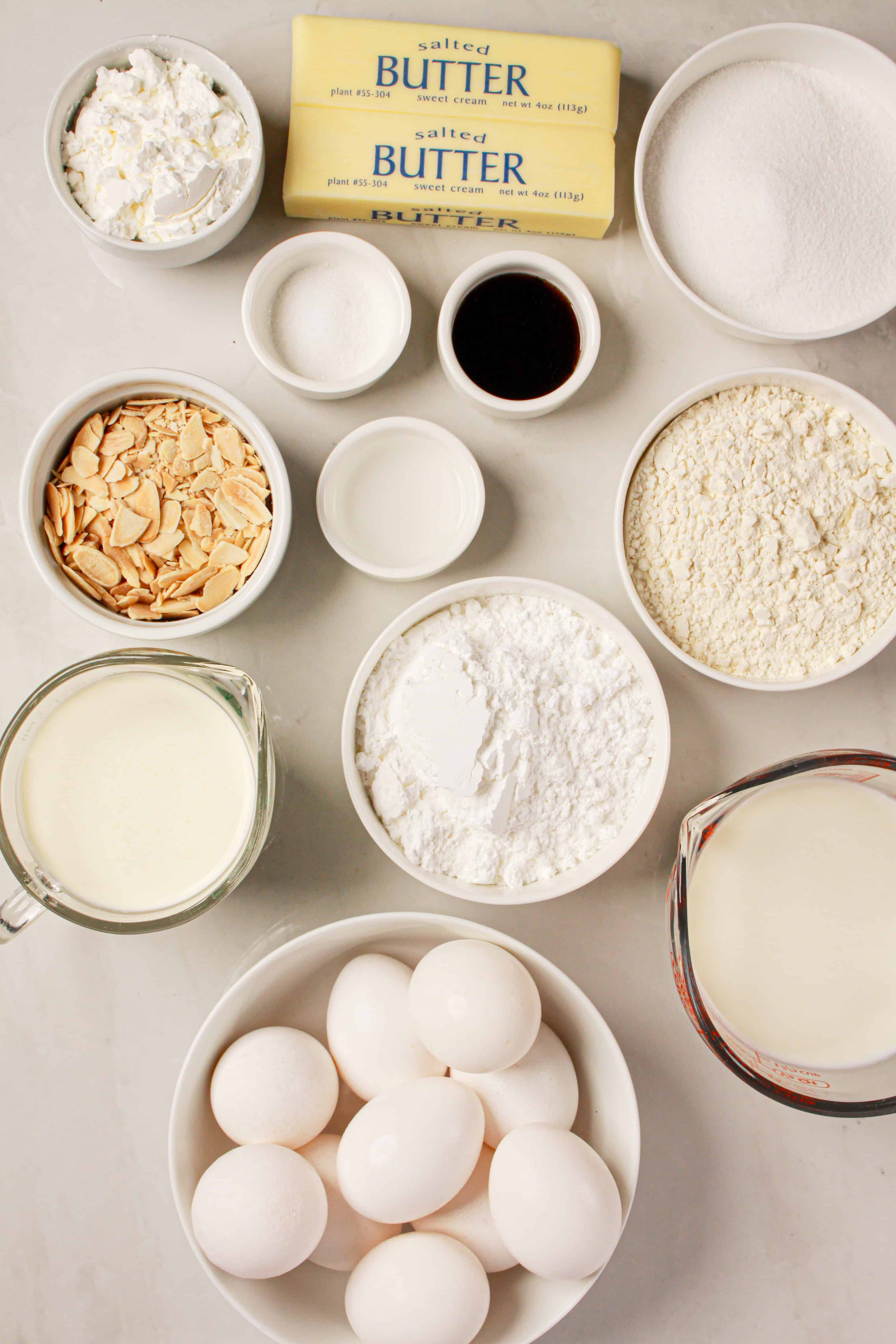 Assorted baking ingredients in bowls, including eggs, butter, flour, sugar, milk, and almonds on a white surface—perfect for creating a Gluten Free Almond Eclair Cake.