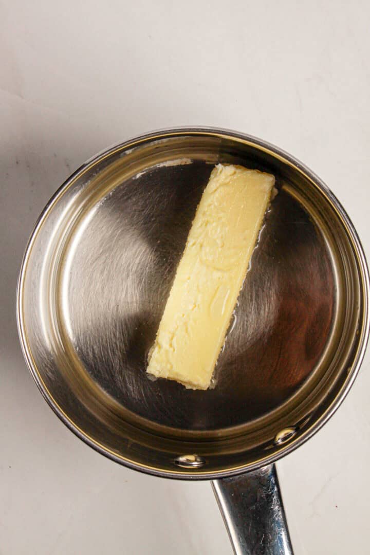A stick of butter in a stainless steel pan on a white surface, perfect for starting your Gluten Free Almond Eclair Cake recipe.