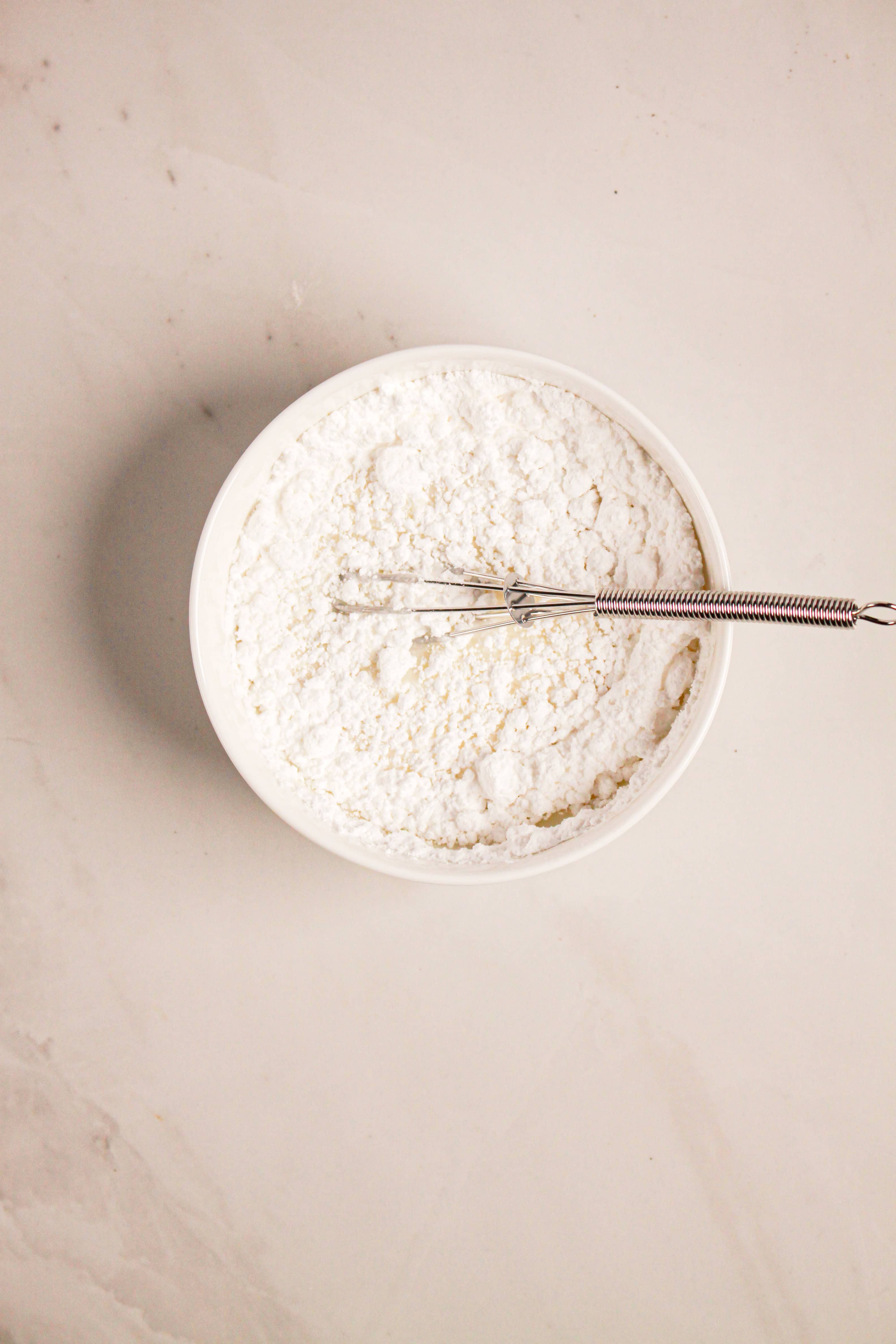 A bowl of white flour with a small whisk on a light-colored surface, ready for creating delicious treats like Gluten Free Almond Eclair Cake.