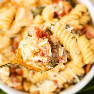 A fork holding cheesy rotini pasta with tomato and herbs above a bowl of pasta.