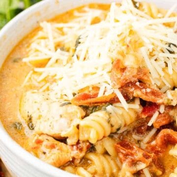 A bowl of creamy pasta with chicken, sun-dried tomatoes, spinach, and shredded cheese on top.