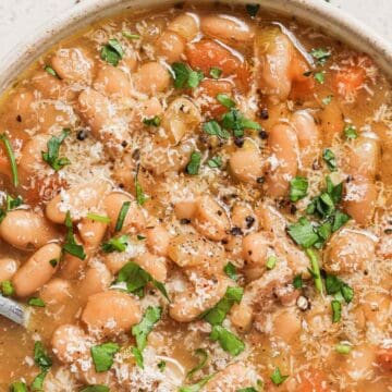 Bowl of white bean soup with chopped parsley, grated cheese, and a spoon on a light surface.