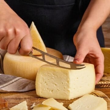 A person slices a block of cheese with a cheese knife on a wooden board, surrounded by wheat and cheese slices—an ideal scene for gifts for cheese lovers.