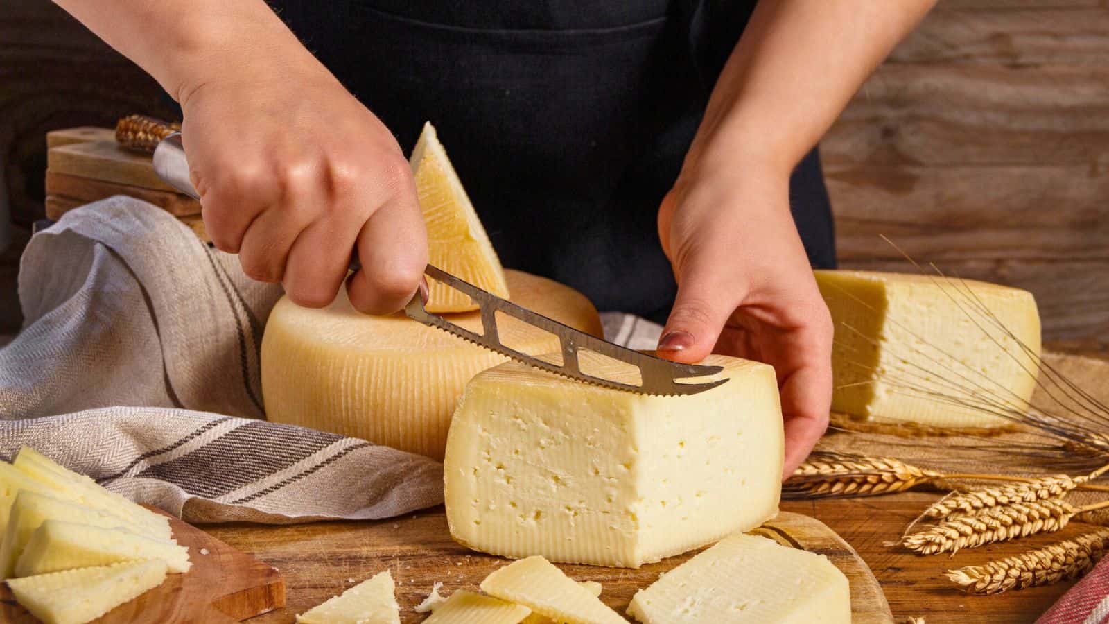 A person slices a block of cheese with a cheese knife on a wooden board, surrounded by wheat and cheese slices—an ideal scene for gifts for cheese lovers.