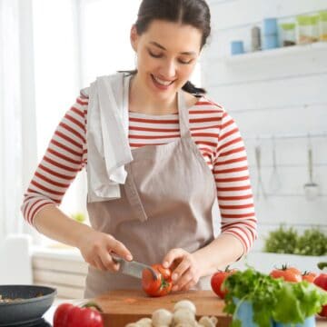 Woman in striped shirt and apron uses essential cooking tools for beginners, slicing a tomato in a bright kitchen with fresh vegetables on the counter.