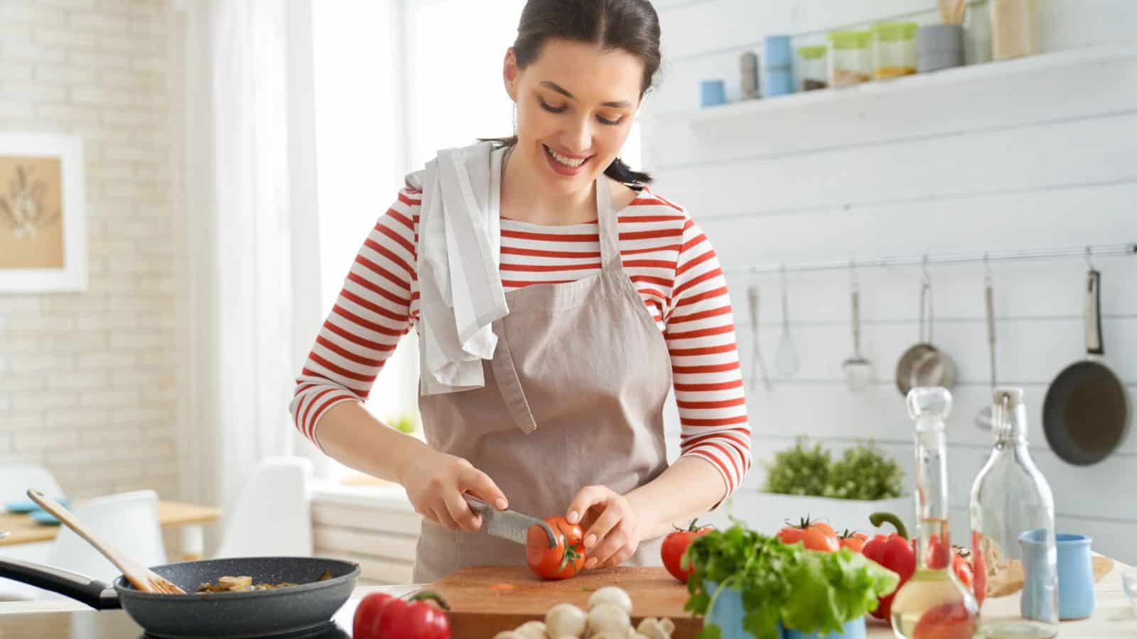 Woman in striped shirt and apron uses essential cooking tools for beginners, slicing a tomato in a bright kitchen with fresh vegetables on the counter.