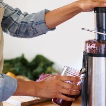 Person in an apron uses a juicer to fill a glass jar with juice in a kitchen.