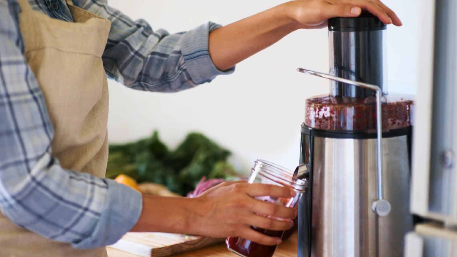 Person in an apron uses a juicer to fill a glass jar with juice in a kitchen.