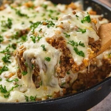 A wooden spoon lifts cheesy, baked casserole topped with melted cheese and fresh parsley from a skillet.