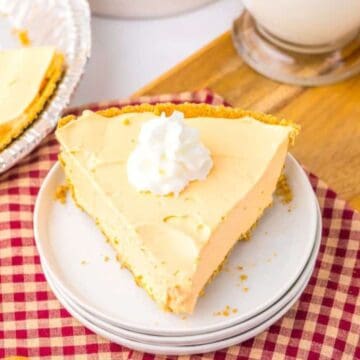 A slice of creamy pie with whipped cream on top sits on stacked plates beside a whole pie, wooden spoons, orange slices, and a glass of milk on a red checkered cloth.