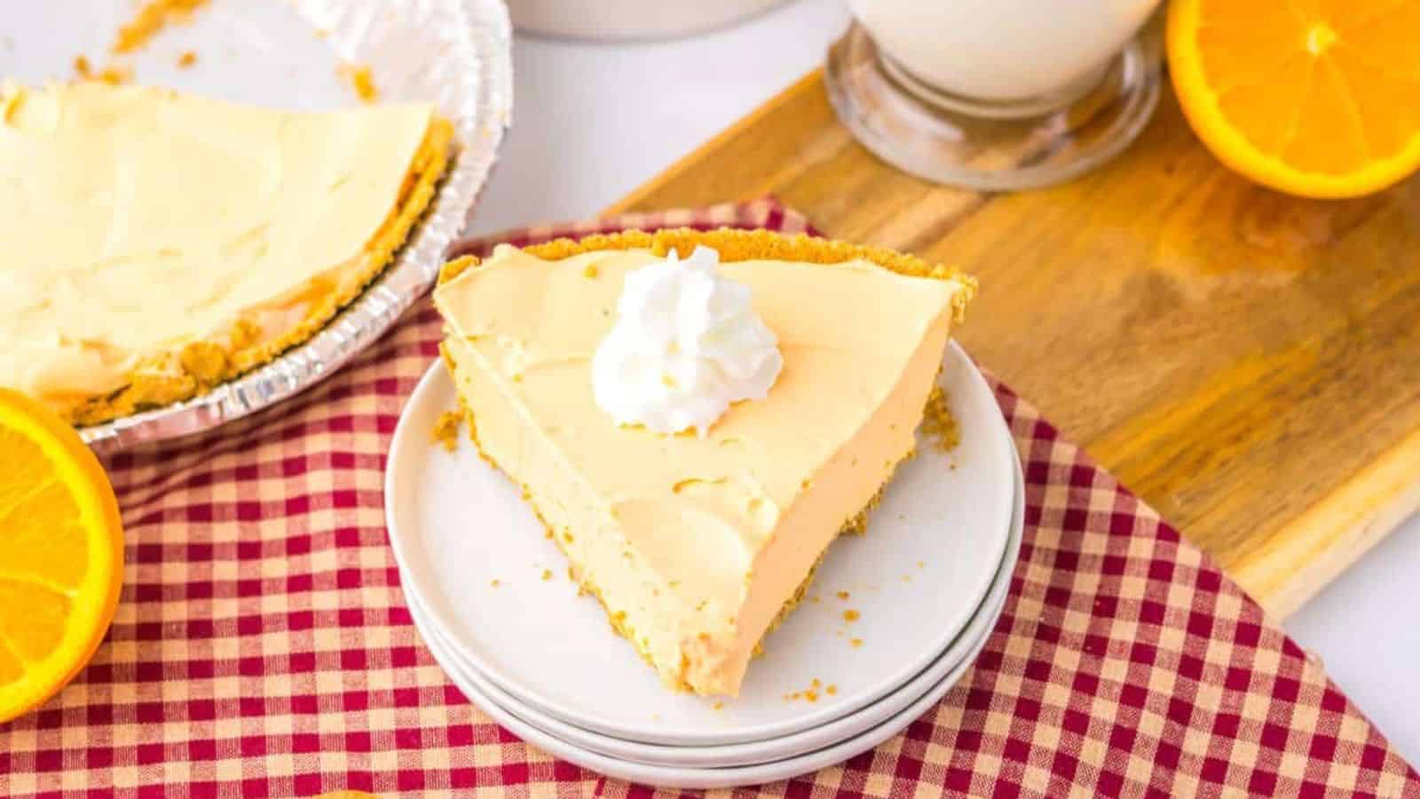 A slice of creamy pie with whipped cream on top sits on stacked plates beside a whole pie, wooden spoons, orange slices, and a glass of milk on a red checkered cloth.