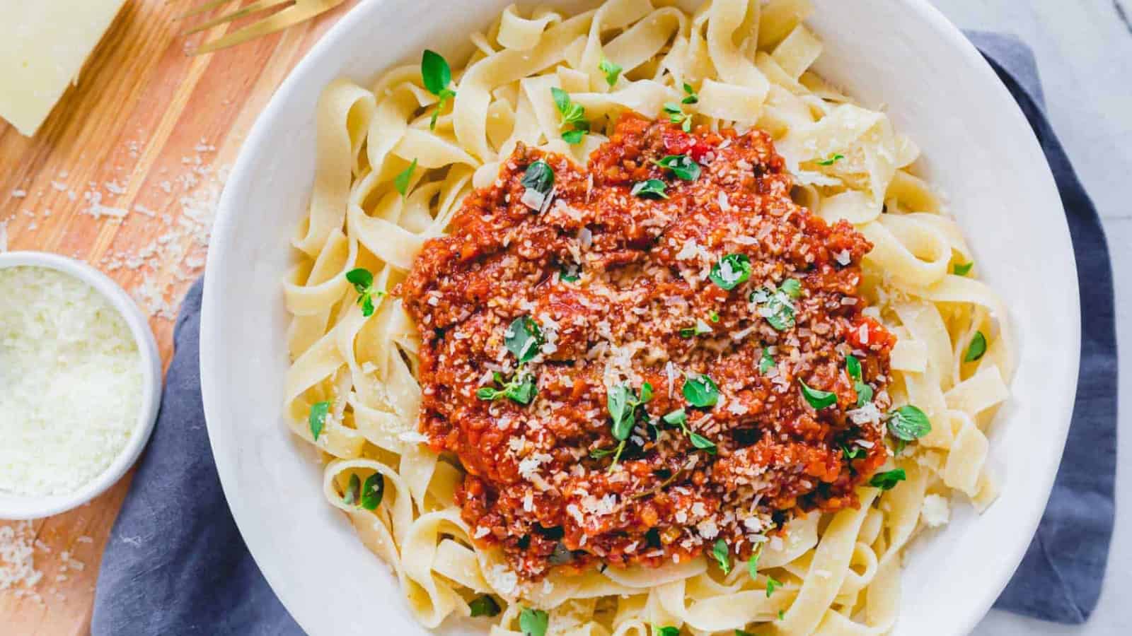 A bowl of fettuccine pasta topped with meat sauce and fresh herbs, with grated cheese on the side.