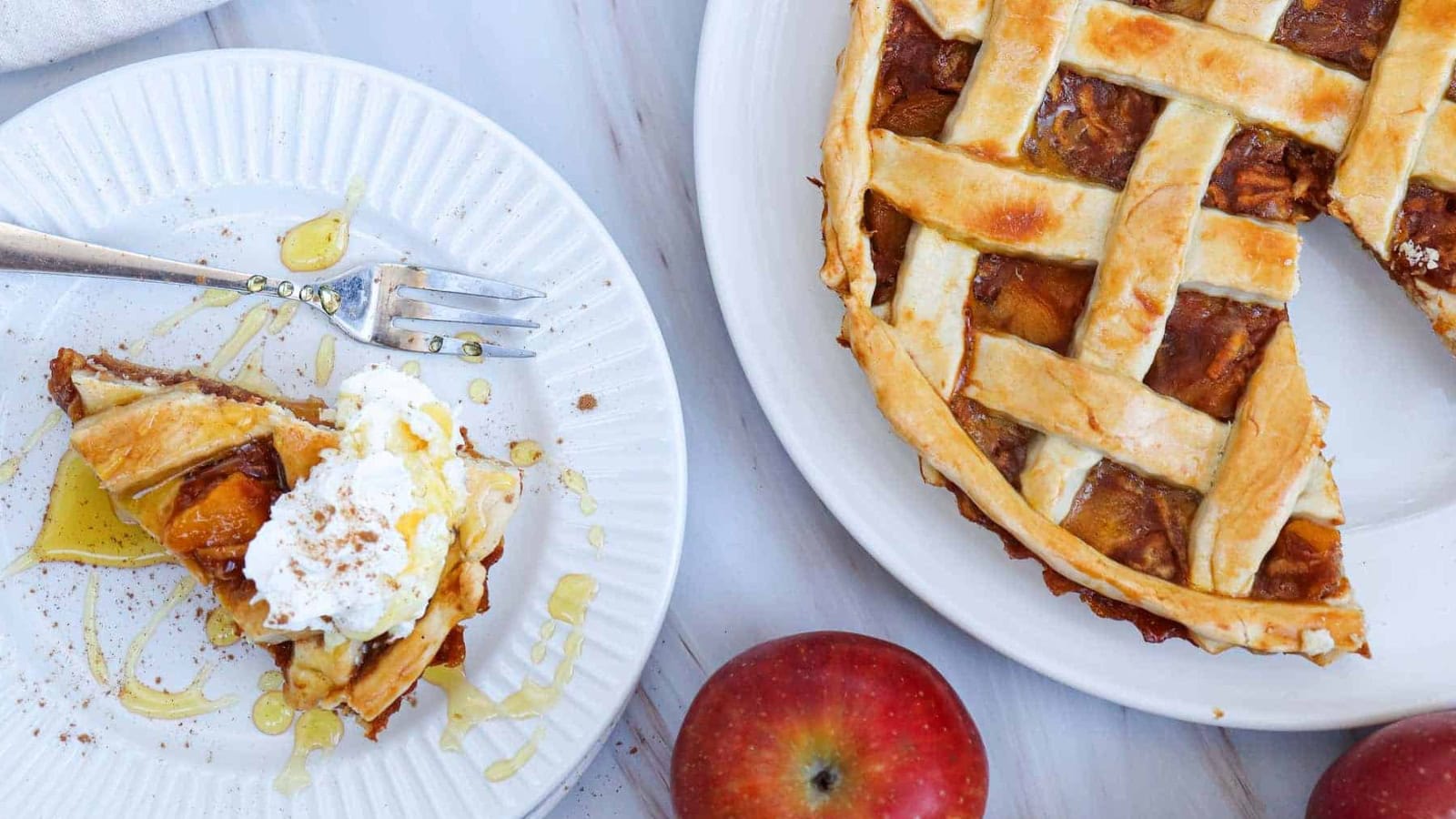 Slice of pumpkin pie with whipped cream and honey on a white plate, with a whole pie and apples in the background.