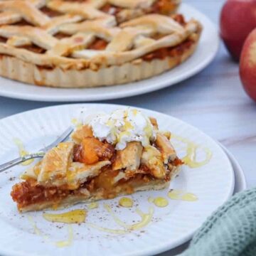 Slice of apple pie with whipped cream and honey on a plate, whole pie and apples in the background.