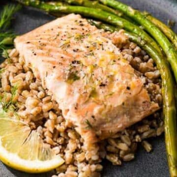 Grilled salmon on a bed of rice with asparagus, a lemon wedge, and fresh dill on a black plate.
