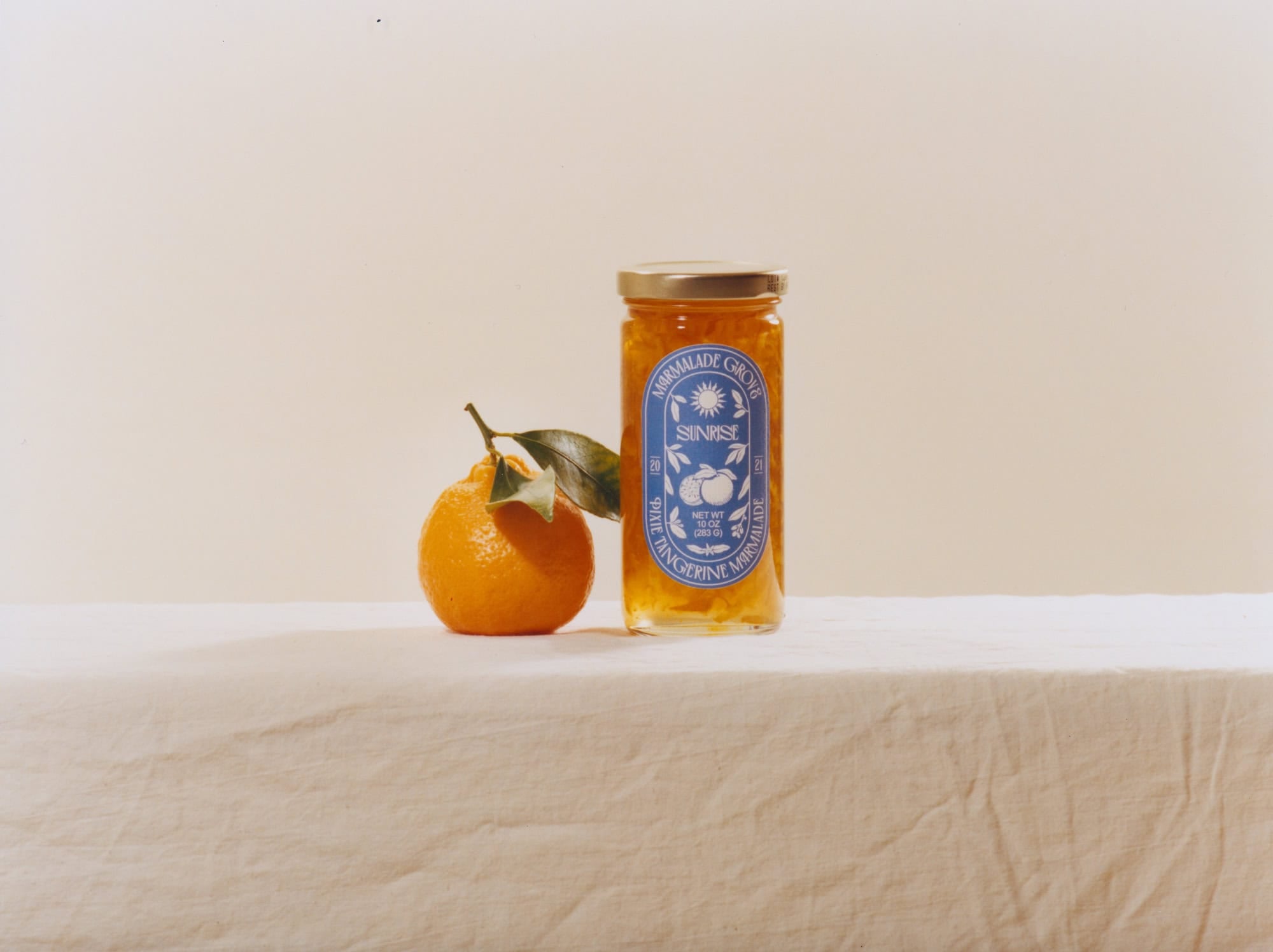 A jar of orange marmalade and a fresh orange with leaves on a white cloth-covered surface—perfect gifts for cheese lovers.