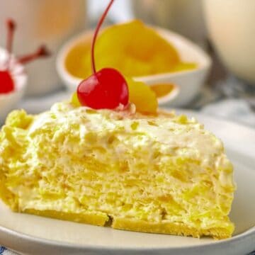 A slice of creamy pineapple pie topped with a cherry on a white plate, with cherries and fruit in the background.