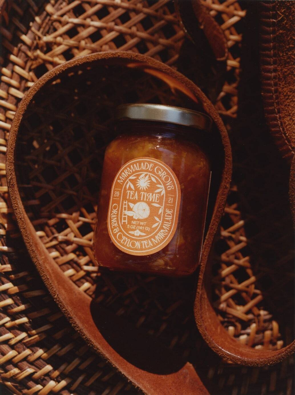 A jar of marmalade sits on a woven surface with leather straps around it, sunlight casting shadows-an ideal touch for gifts for coffee and tea lovers.