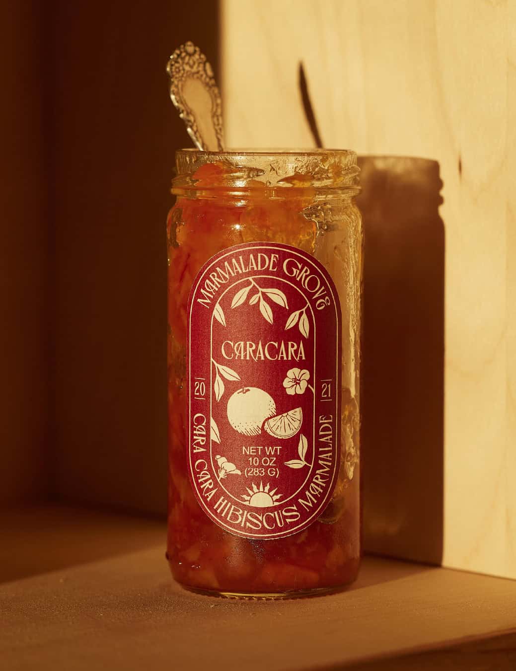 A jar of Cara Cara hibiscus marmalade with a spoon inside, sitting in warm sunlight against a wooden background-perfect as gifts for cheese lovers.