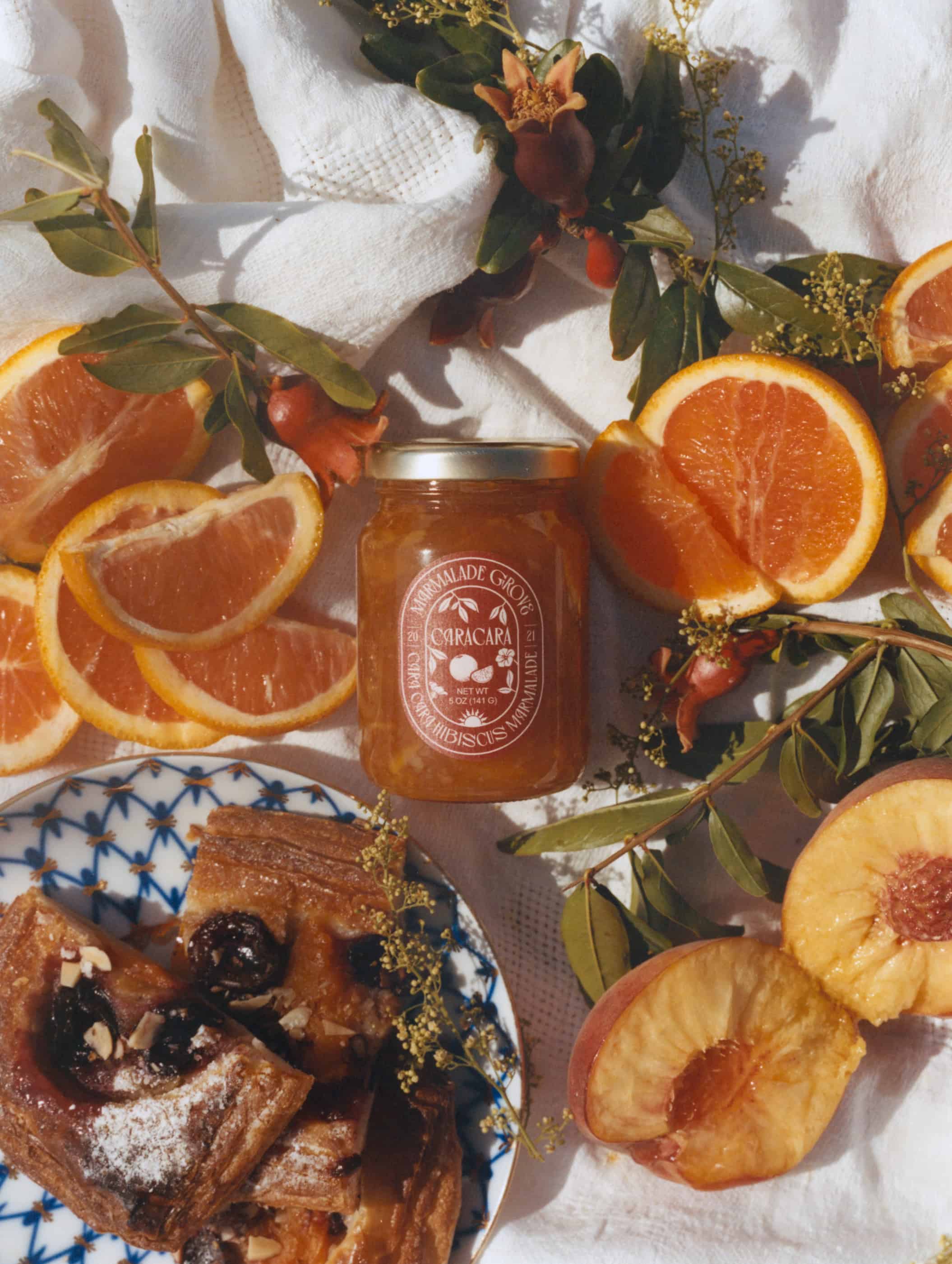 A jar of orange marmalade surrounded by sliced oranges, peaches, and pastries on a white cloth-perfect as gifts for cheese lovers.