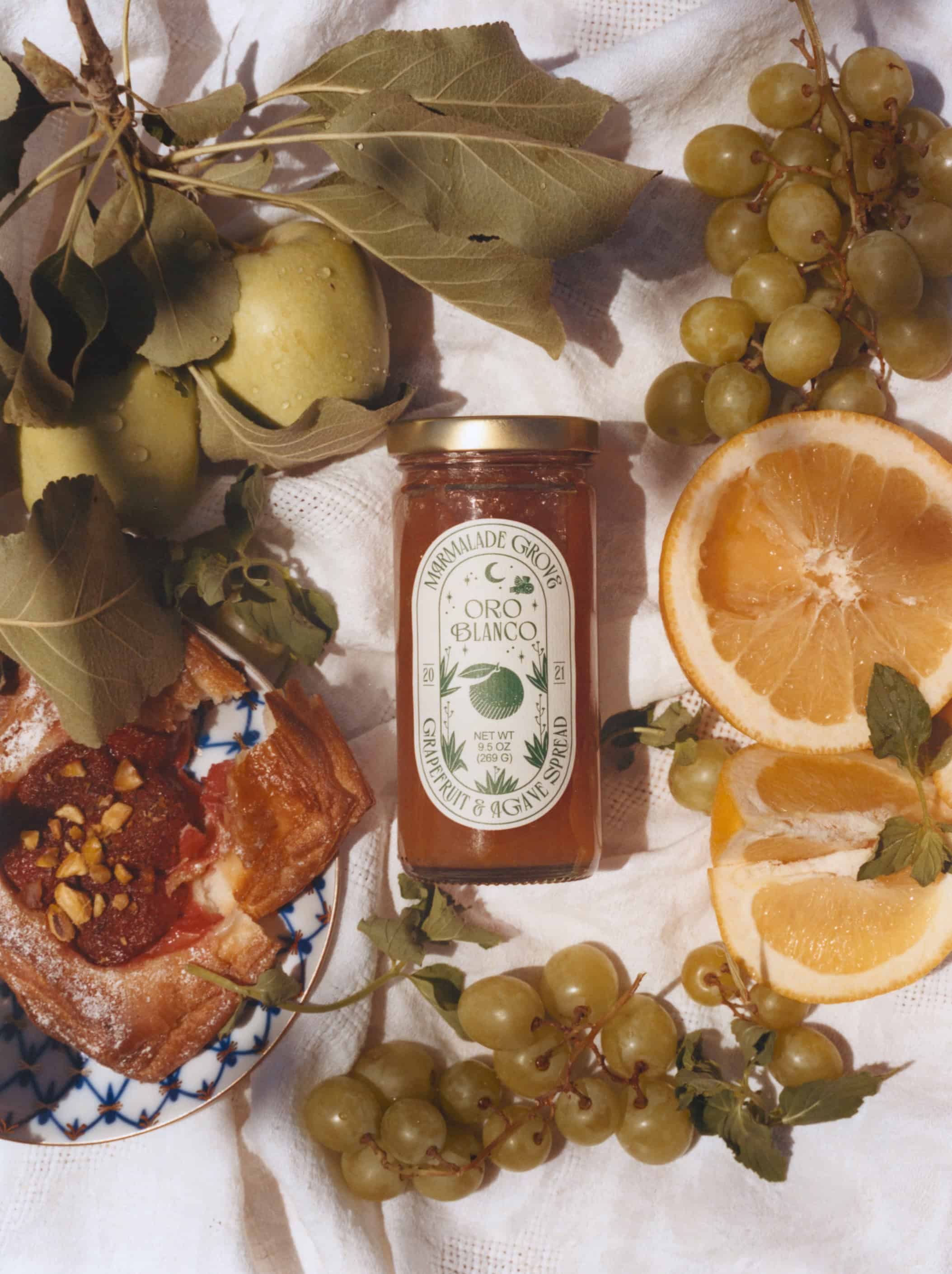 Jar of honey surrounded by green grapes, apples, orange slices, and pastry on a white cloth—perfect gifts for cheese lovers seeking a delightful pairing.