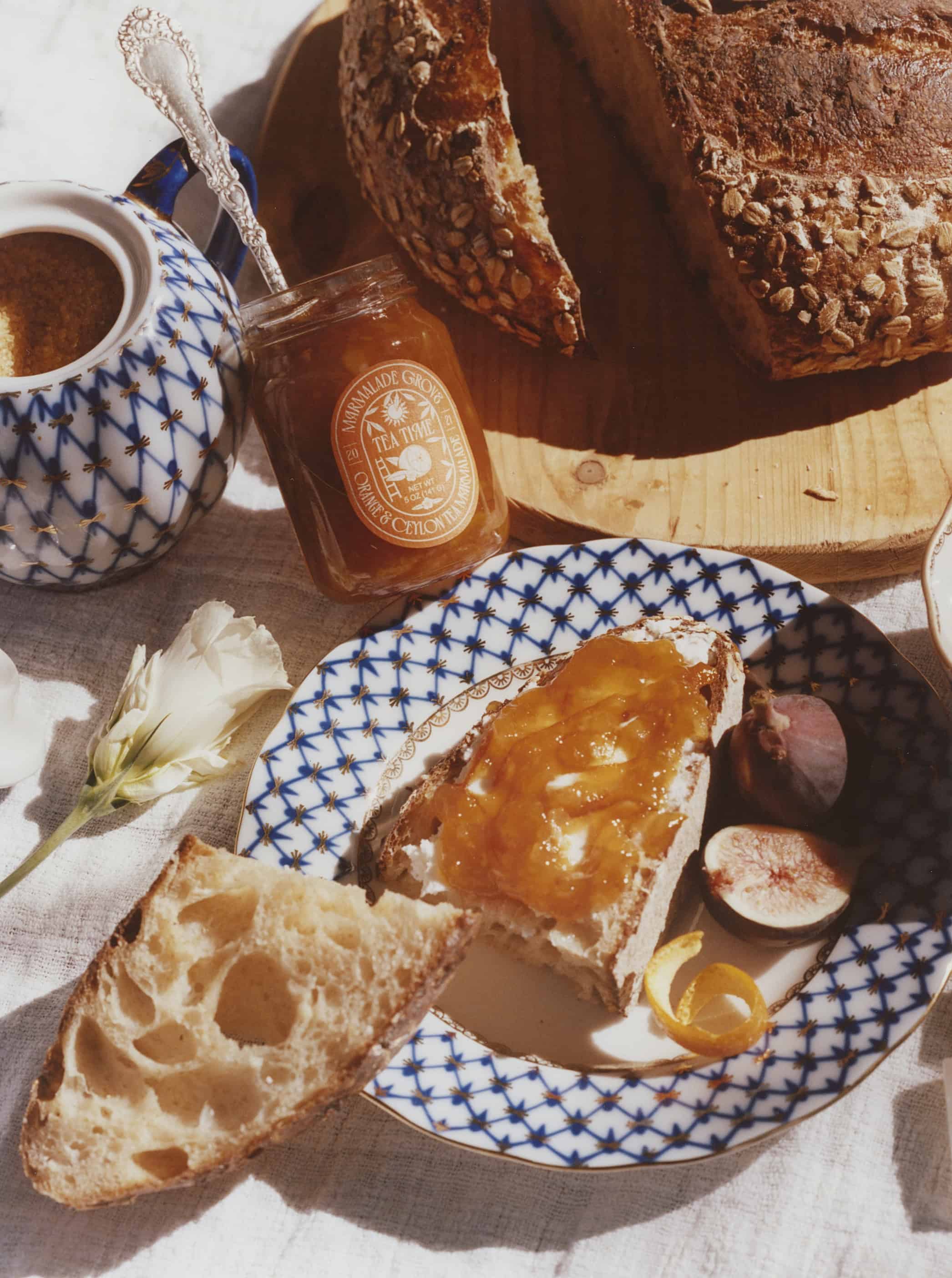 A slice of bread with marmalade, figs, and orange peel on a patterned plate, with bread and a marmalade jar nearby-perfect gifts for coffee and tea lovers who appreciate artisanal flavors with their morning brew.