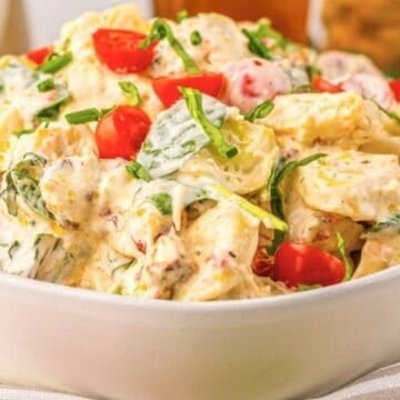 A bowl of creamy potato salad with cherry tomatoes, green onions, and herbs on a striped cloth.